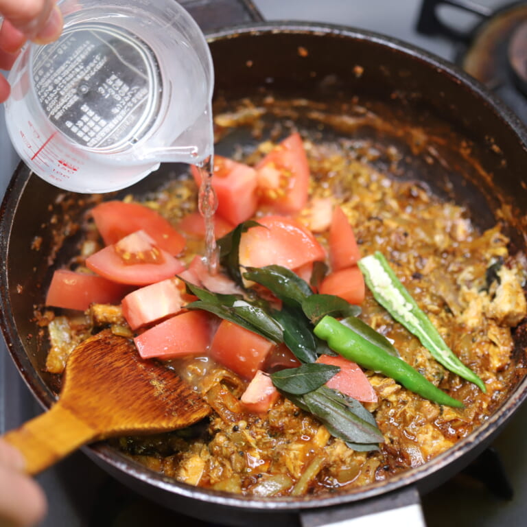 手順 5: トマトを加えざっくりと炒め合わせたら少しずつ水を加えカレーリーフ 、青唐辛子を加え蓋をして弱火で１０分煮込む。