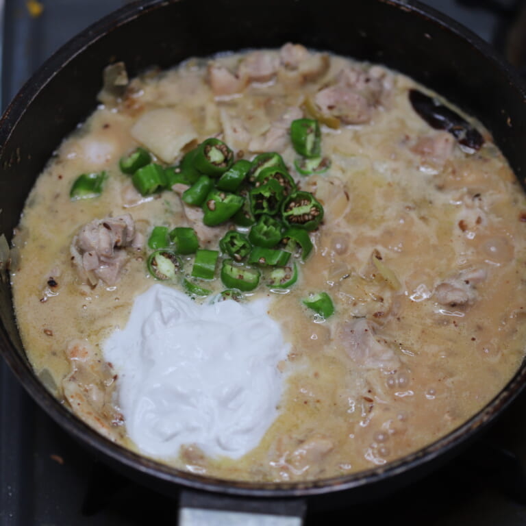 手順 5: 水を少しずつ加えて伸ばし、蓋をして１５分ほど煮込む。鶏もも肉に火が通ったらココナッツミルク、ししとうを加え、一煮立ちさせ